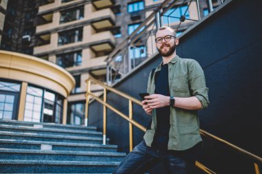 App ücretsiz genel internet kullanarak modern smartphone cep telefonu ile güncelleştirilirken kameraya gülümseyen pozitif sakallı genç adam portresi. Hücresel ayakta merdivenlerde rahat kıyafet bloglama neşeli hipster adam