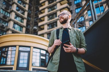 Aşağıda görünümü durulması sakallı öğrencinin elinde smartphone ile yürüyüş sırasında şehir zevk. Düşünceli hipster adam uzağa bakarak ve modern telefon tutarak gülümseyen gözlük rahat giyinmiş