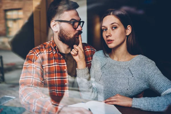 Half length portrait of attractive young woman making boyfriend stop talking during conflict in relationship.Casual dressed girlfriend told secret to bearded man and showing silence