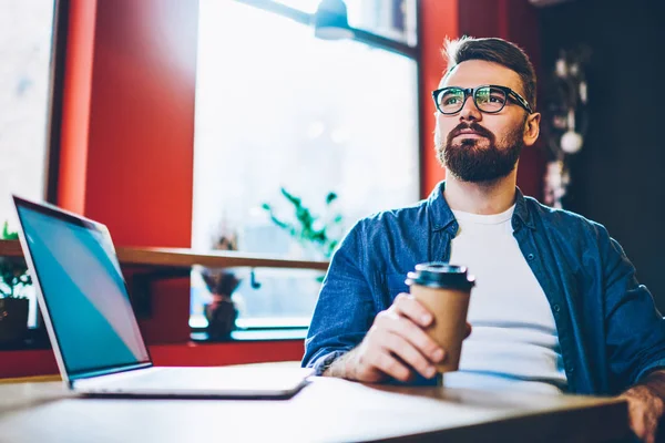 Dreamy bearded freelancer in eyewear for vision correction pondering on business strategy of developing own startup during coffee break.Pondered hipster guy thinking on idea for blog sitting at laptop