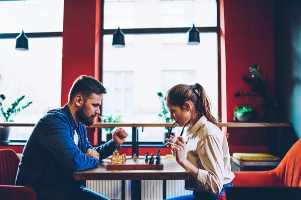 Side view of concentrated two hipsters thinking on tactical strategy of next move on chessboard during sport competitions in chess.Young couple in love playing in favourite game in free time
