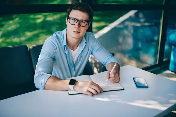 Portrait of smart student in eyeglasses for vision correction looking at camera while working on writing homework in notebook.Successful young man making notes of startup project sitting at table