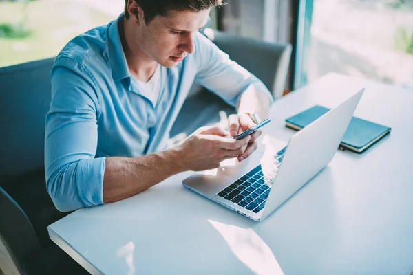 Młody Człowiek Freelancer Sprawdzania Powiadomień Smartfonie Pracując Zdalnie Netbook Przy — Zdjęcie stockowe