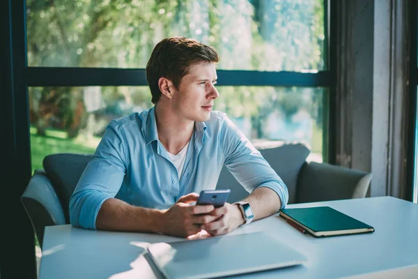 Pondering young man looking out of window while typing text message on modern smartphone device and installing app using free 4G internet.Thoughtful hipster blogger updating profile on telephone