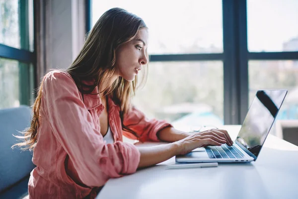 Side view of concentrated student attentively reading information from website on modern laptop computer using wireless internet,Pensive young woman keyboarding text for course work on netbook