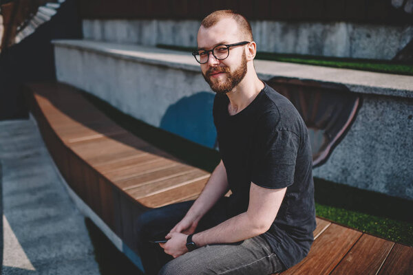 Portrait of caucasian bearded young man in eyeglasses for vision correction smiling at camera while sitting in urban setting in city.Positive hipster guy in eyewear spending free time on street