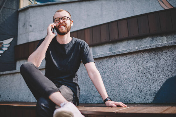 Portrait of cheerful young man in eyeglasses with beard smiling at camera while communicating with friend in roaming on smartphone device.Positive hipster blogger calling on telephone device