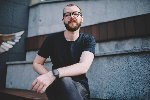 Portrait of cheerful bearded young man dressed in stylish casual wear smiling at camera while spending free time in urban setting.Positive hipster guy in eyewear sitting outdoors in centre of city
