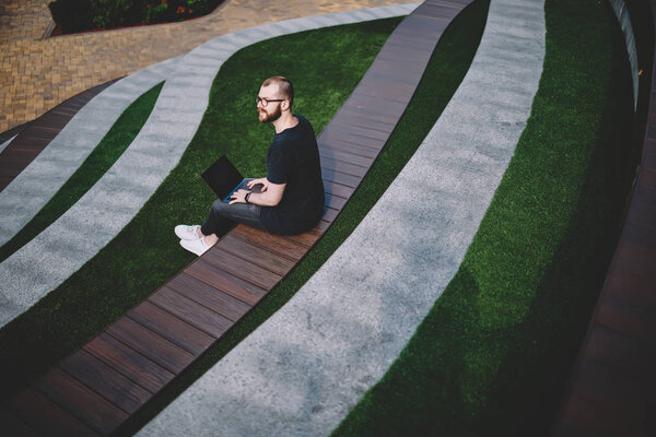 Pondering young man looking away while searching information on website on modern laptop computer sitting outdoors in park.Thoughtful hipster freelancer thinking on creative ideas for publication