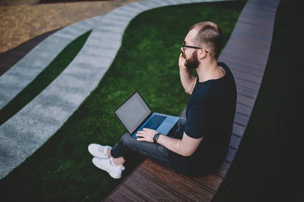 Caucasian hipster guy updating software on modern laptop computer using 4G internet connection in national park.Young man installing app for remote work on netbook device in urban setting