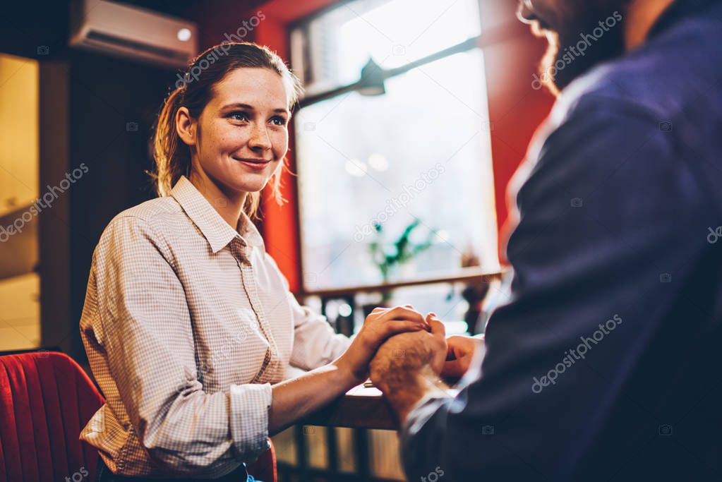 Charming young woman discussing future plans on married life on romantic date in cafeteria.Happy hipster girl listening attentively boyfriend holding hands each other sitting at meeting table