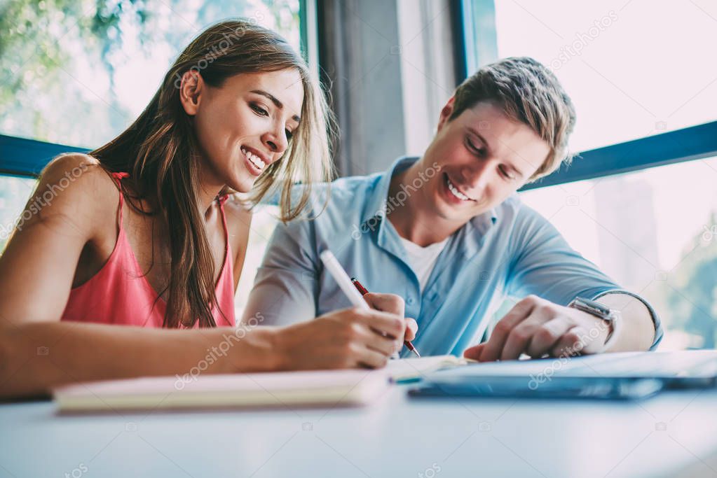 Cheerful young man and woman laughing while preparing for upcoming exams together in coworking.Successful students collaborating on common project making notes of creative ideas in notepad