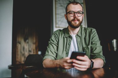 Kafede ücretsiz Wi-Fi kullanarak modern smartphone içinde online sohbet ederken kameraya bakarak gözlük sakallı genç adam portresi. Hipster blogger cep internet bağlantısı üzerinden App yükleme