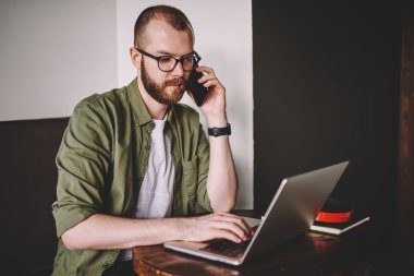 Dalgın smartphone aygıtında çağrılırken dizüstü bilgisayarda posta kontrol uzmanı. Sırasında cep telefonuyla konuşan genç adam sakallı kablosuz internet kullanarak modern ağ dizüstü bilgisayarı, uzaktan çalışma