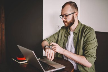 Dalgın sakallı genç smartwatch denetimi zamanında sırasında kafede dizüstü bilgisayarda çalışan adam. Konsantre hipster öğrenci sınav netbook için hazırlarken marka kol saati yönetme