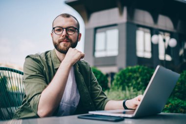 Gözlük mesafesi iş modern dizüstü bilgisayar başında otururken kameraya bakarak sakallı öğrenci portresi. Serbest meslek sahibi netbook aygıt açık havada kafede çalışan genç adam
