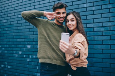 Genç erkek ve kadın giyim fotoğrafı çekmek modern cep telefonu kullanarak kamera bakarak içinde smartphone kamera birlikte, ayakta üzerinde funny fotoğraf için poz neşeli hipster çift portresi