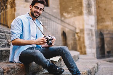 Hipster adam turistik Tur, resimler yapmak için ekipman kontrol kamera bakarak gözlük olumlu erkek fotoğrafçı sırasında dinlenme merdivenlerde oturan vintage fotoğraf makinesi ile gülümseyen portresi