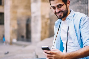 Neşeli hipster adam açık havada oturma, sosyal ağlar üzerinden cep doğrulayan yeni arkadaşıyla Erkek seyahat blogger memnun gülümseyen smartphone arkadaş gelir mesaj kontrol gözlükler
