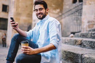 Gitmek için mutlu erkek moda gözlük cep kullanarak şehirde serbest zaman harcama'sosyal ağlar ile kahve Sokağı'nda oturan sohbet için smartphone neşeli hipster adam portresi