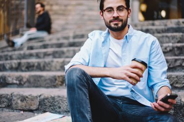 Hücresel tutan merdiven ve kahve fincanı, Genç hipster adam cep telefonu kullanarak şehirde bahar gününde sokak eğlence üzerinde rahat kıyafet harcama zaman içinde oturan gözlük yakışıklı erkek portresi