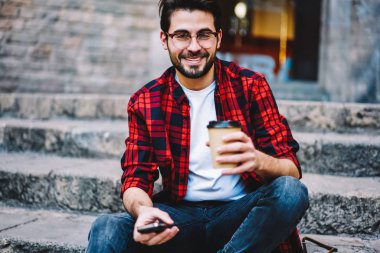 Yakışıklı erkek giyim ve gözlük açık havada dinlenme ile kahve Kupası, neşeli hipster adam sokak holding smartphone üzerinde oturan kamera seyir gitmek portresi bağlı 4 g için Internet