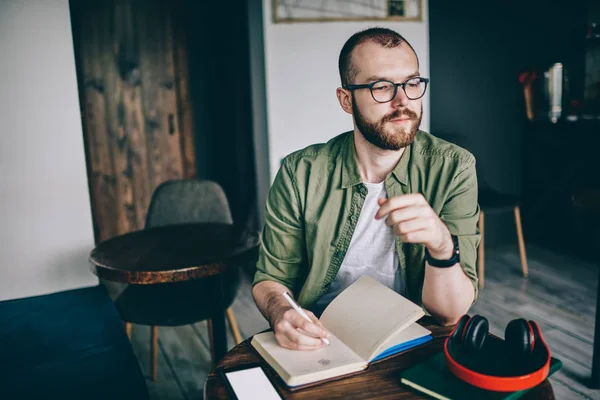 体贴胡子的年轻男子在眼镜的创意 在咖啡桌上坐在咖啡馆的笔记与记事本的想法 在咖啡馆里做家庭作业的时髦男人的思考 — 图库照片
