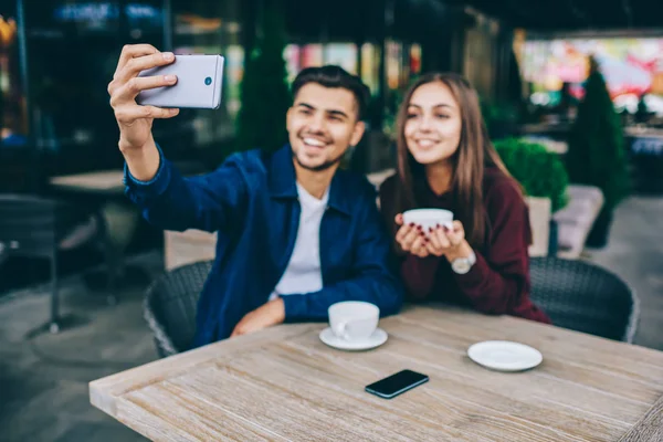 Cheerful male and female hipsters posing for selfie on smartphone camera at cafe terrace during coffee break, selective focus on modern mobile phone in hand of millennial man taking picture with friend