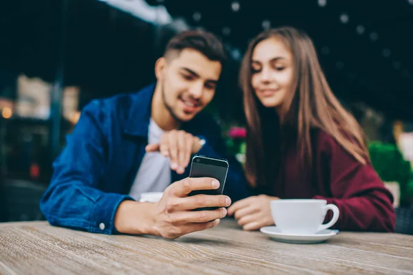 Selective focus on smartphone in hand of hipster guy using gadget for showing multimedia files to hi girlfriend on free time,couple silhouette on blurred background watching video on telephone