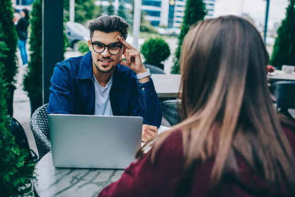 Portrait of confused bearded guy looking at camera streets during conversation with female colleague, back view of woman talking to hipster guy in spectacles sitting with laptop computer on terrace