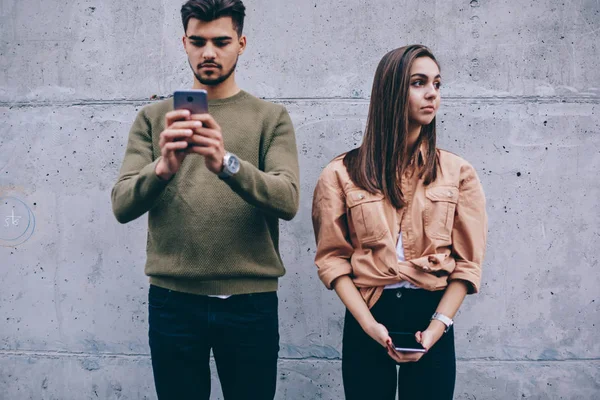 Young sad woman looking away while her boyfriend concentrating on online communication in social networks standing on wall,man holding cellular addicted to internet and gadgets ignore conversation