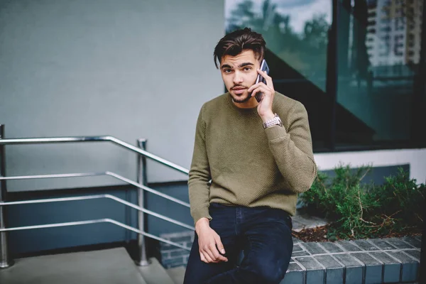 Portrait of serious bearded man having mobile phone conversation with service operator, handsome hipster guy sitting outdoors making telephone call in roaming for communication via telephone