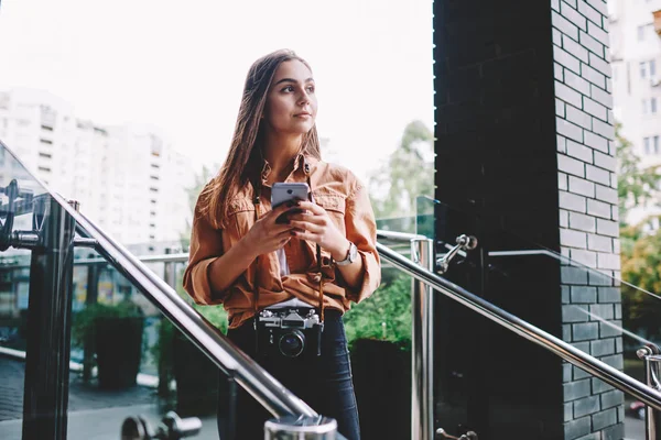 Pensive hipster girl looking away holding mobile phone connected to 4G internet standing outdoors with vintage camera, woman amateur using application for networking  standing on urban setting