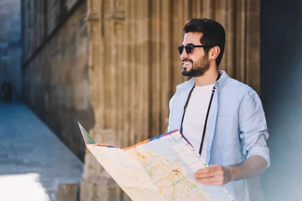 Cheerful bearded hipster guy satisfied with sightseeing tour in town on summer weekends,happy male traveler holding map for navigate in city and get to destination standing on street in sunglasses