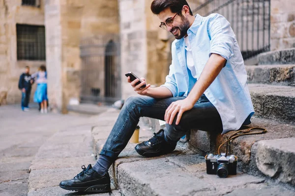 Happy hipster guy laughing at funny video from social network browsed on cellular connected to 4G internet, positive male traveler blogging via smartphone enjoying connection in roaming outdoors