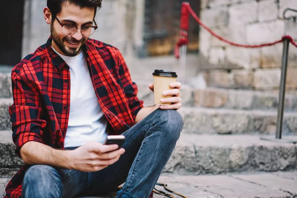 Cheerful hipster guy chatting with followers in social networks updating feeds on cellular sitting on street, male tourist satisfied with mobile internet in roaming sending text messages on telephone