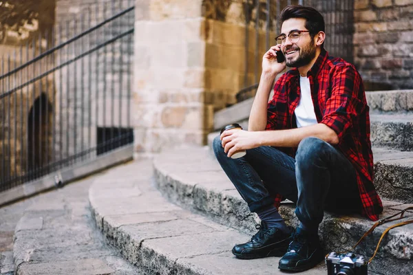 Cheerful hipster guy in spectacles making telephone call in roaming satisfied with good connection, male tourist using international tariffs for mobile phone conversations sitting on street