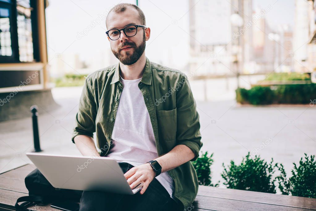 Portrait of bearded young man in eyeglasses looking at camera while working on freelance at laptop computer using public 4G internet.Hipster guy sitting on street with netbook during distance job