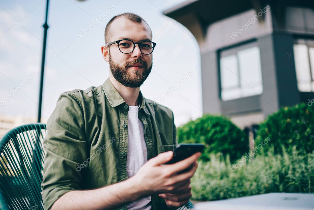 Portrait of bearded young man in eyeglasses holding telephone in hands and updating app using internet connection.Casual dressed hipster guy installing programme on smartphone while looking at camera