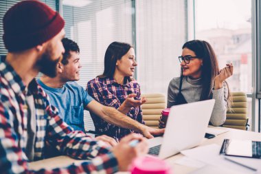 Hipster öğrencilerin birbirleriyle iletişim kurmak ve işbirliği tablo ofiste dizüstü bilgisayar ile Toplantı oturma planını tartışıyor rahat kıyafet. Takım görev üzerinde işbirliği gençlerin