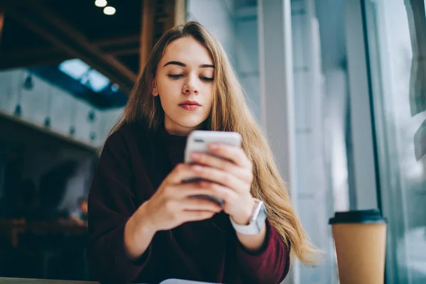 Attractive hipster girl waiting for call on smartphone while sitting indoors and connected to wifi zone, charming female student chatting with friends using mobile phone with 4g internet at cafe