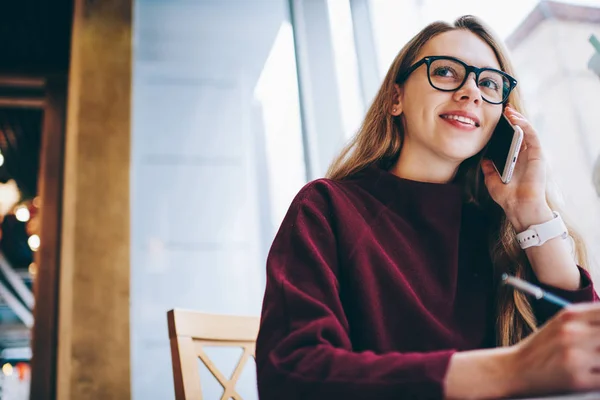 Positive hipster girl calling to customer service using application on mobile phone while sitting at university library, successful female journalist wear in spectacles using technology indoors