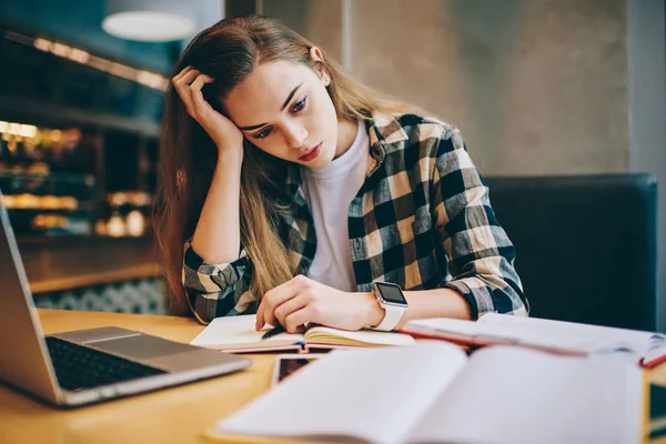 Tired female student preparing for important exam trying find answers for questions using modern laptop and books indoors, concentrated woman feeling headache from course work at university library