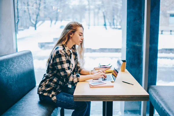 Charming female copywriter typing email while downloading new file on modern laptop sitting near window indoors, good looking hipster girl 20s searching information for course work via netboo