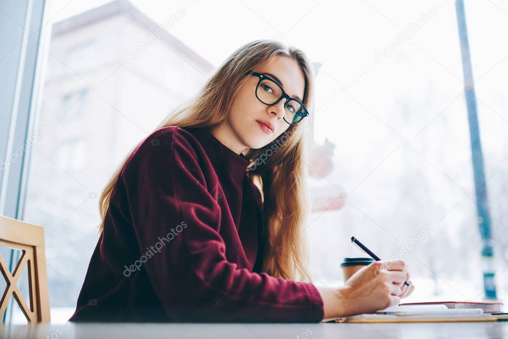 Portrait of serious female student sitting at table at coffee shop and studying, beautiful hipster girl looking at camera in break of learning indoors, charming woman writing organisation plan