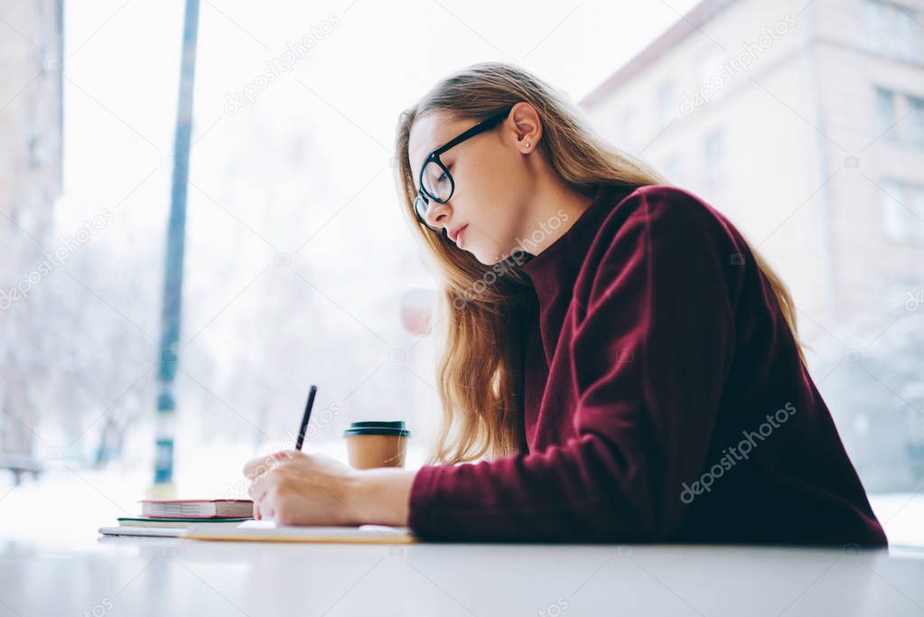 Concentrated young clever writer wear in trendy eyeglasses creating new articles while sitting at coffee shop, pondering female student preparing for exam thinking on answers for question indoors