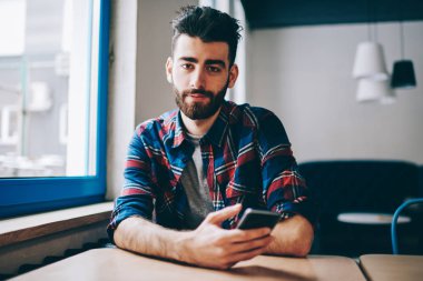 Yakışıklı erkek Blogger uzay smartphone ile el, coworking oturma portre sakallı kameraya bakarak ve sohbet için cep telefonu wifi için bağlı tutan beyaz hipster adam