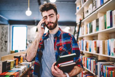 Üniversite kampüsünde, rahat telefon görüşmesi yaparken kameraya bakarak iyi görünümlü hipster adam erkek öğrenci sınava hazırlanıyor ve smartphone üzerinden arkadaşıyla konuşmak için kitap seçme giyinmiş