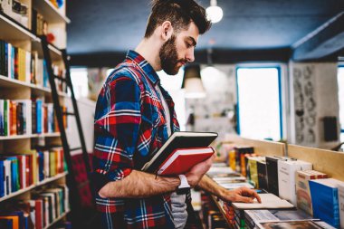 Yakışıklı şık hipster adam üniversite kütüphanesi ders kitabı listeden seçerek literatürde, iyi görünüyor sakallı adam otodidakt ve kapalı eğitim Kitabevi düşünmeye romanı okuma