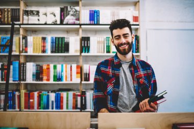 Half length portrait of smiling bearded male teenager selecting book in library of college standing near shelves, happy successful hipster guy picking literature and making research in bookstore clipart